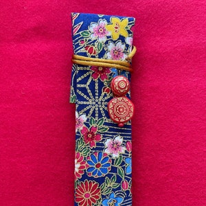 May include: A blue fabric pencil case with a floral pattern and gold accents. The case has a brown cord with two red buttons.