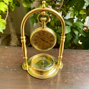 May include: A gold-toned decorative stand with a compass and a pocket watch hanging from it. The watch face has Roman numerals and the text "Victoria London 1876".