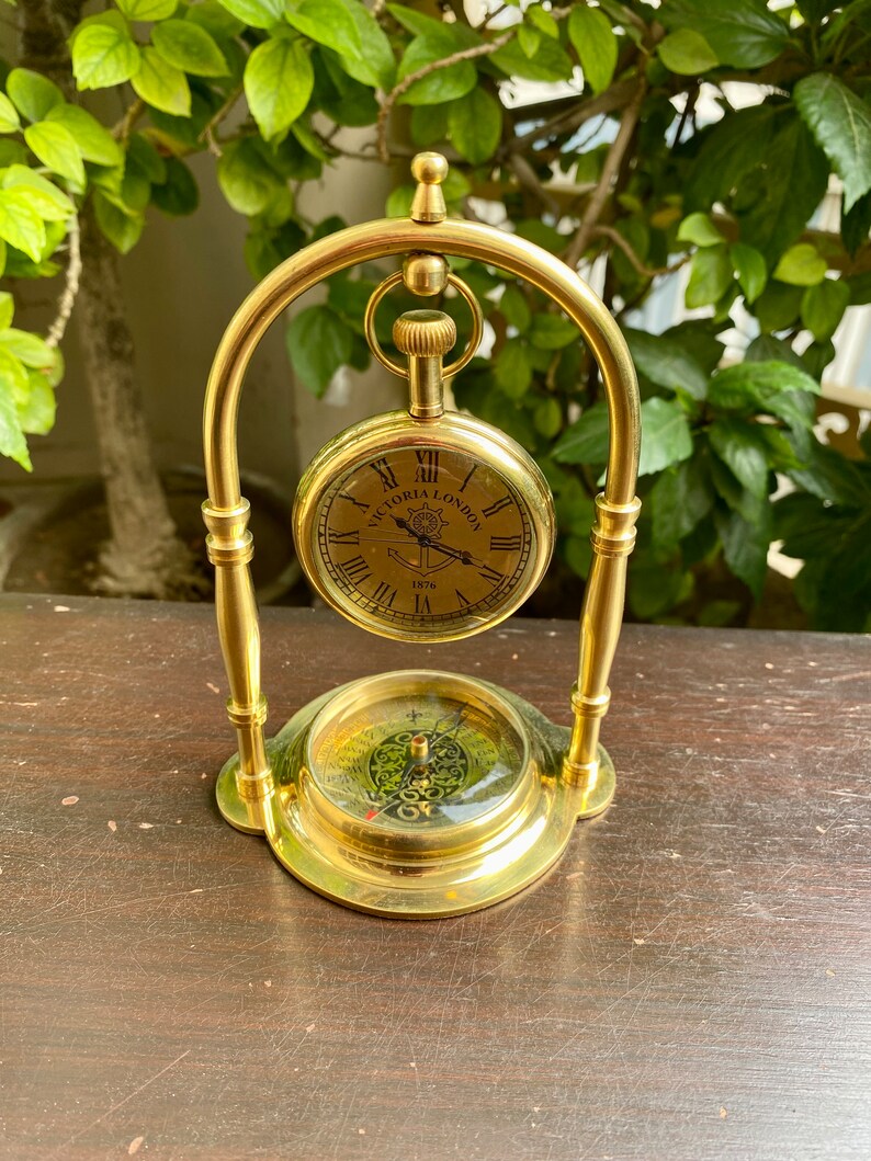 May include: A gold-toned decorative stand with a compass and a pocket watch hanging from it. The watch face has Roman numerals and the text "Victoria London 1876".