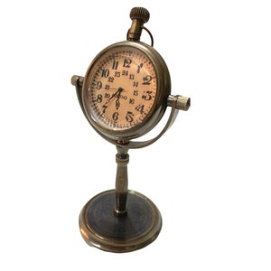 May include: A brass desk clock with a round face and Roman numeral markings. The clock has a stand with a round base and a decorative design.
