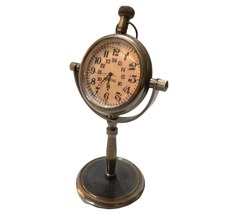 May include: A brass desk clock with a round face and Roman numeral markings. The clock has a stand with a round base and a decorative design.