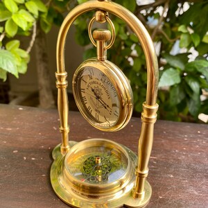 May include: A gold-toned pocket watch with a compass on a stand. The watch face has Roman numerals and says "Victoria London 1876".
