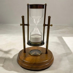 May include: A brass hourglass with a wooden base and a compass design on the top. The hourglass is filled with white sand and has a clear glass bulb.