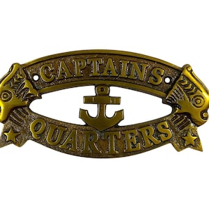Pode incluir: Placa de metal cor bronze com as palavras "CAPTAINS QUARTERS" em letras em relevo. Uma âncora está centrada no meio. A placa tem detalhes decorativos de peixes e estrelas em cada extremidade.