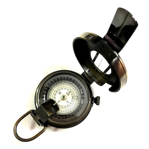 K&ouml;nnte beinhalten: Ein Vintage-Kompass aus Messing mit wei&szlig;em Zifferblatt und schwarzen Markierungen. Der Kompass hat einen Klappdeckel und eine Schlaufe zum Anbringen an einem G&uuml;rtel oder Rucksack.