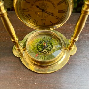 May include: A gold-toned clock and compass set with a round clock face that reads "VICTORIA LONDON 1876". The compass face has a black and gold design with the cardinal directions and points of the compass.