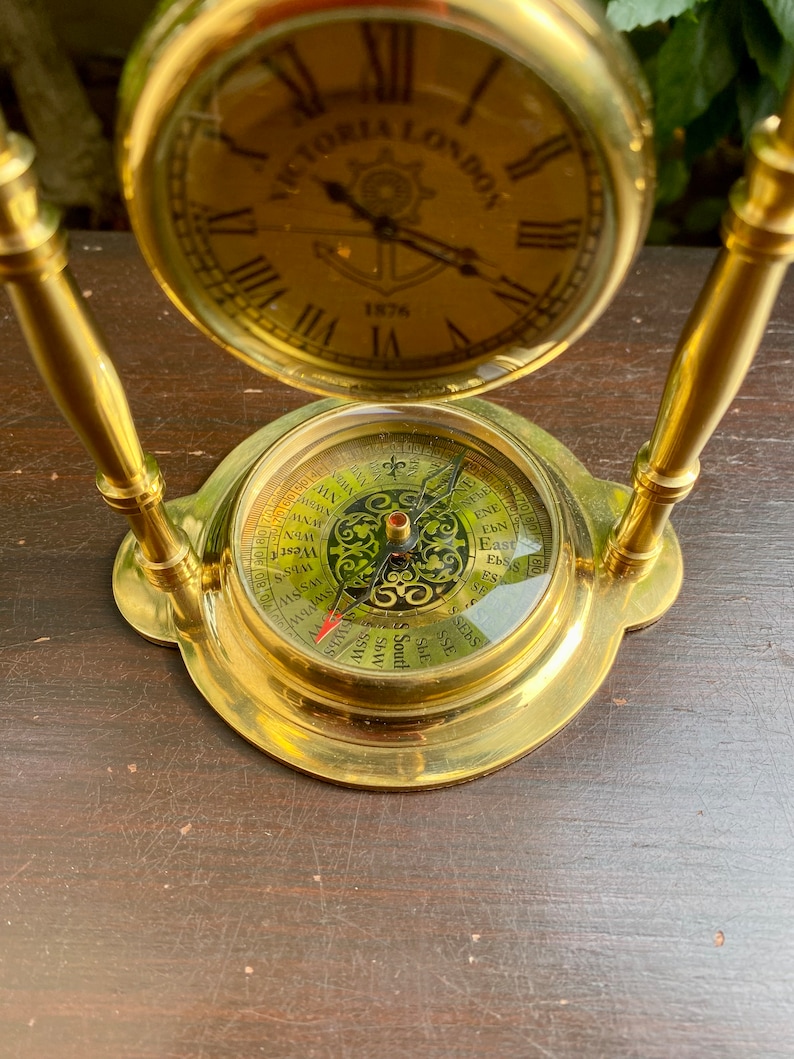 May include: A gold-toned clock and compass set with a round clock face that reads "VICTORIA LONDON 1876". The compass face has a black and gold design with the cardinal directions and points of the compass.