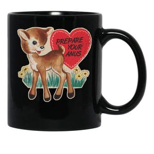 May include: Black ceramic mug featuring a cartoon deer with a red heart that reads "Prepare Your Anus." The deer is brown and white, with yellow flowers and green grass. The mug has a black handle.