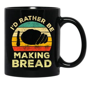 May include: Black ceramic coffee mug with the text "I'd Rather Be Making Bread." The design features a bread loaf illustration with a retro sunset color scheme of teal, yellow, orange, and red.