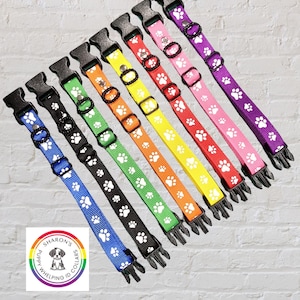 May include: A collection of colorful dog collars in various colors, including blue, black, green, orange, yellow, red, pink, and purple. Each collar features a repeating pattern of white paw prints and a black plastic buckle. The collars are for puppies.