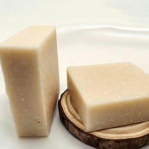 May include: Two bars of handmade soap, one on a wooden stand. The soap is a light beige color and has a grainy texture.