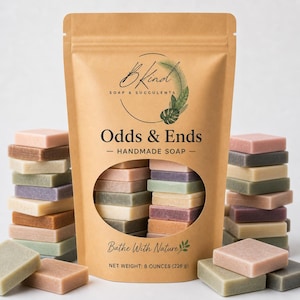 May include: A kraft paper bag labeled "BKind Soap & Succulents" with "Odds & Ends - Handmade Soap" printed on it. The bag contains various small, square soaps in shades of pink, green, and brown. Stacks of similar soaps are arranged around the bag.