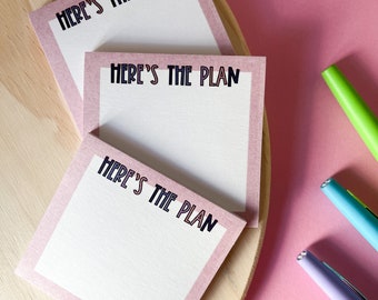 May include: Three pink notepads with the text "Here's the plan" in black lettering. The notepads are on a wooden surface with two pens on a pink background.