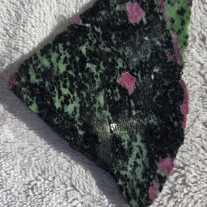 May include: A green and pink gemstone with black speckles. The gemstone is irregularly shaped and has a smooth, polished surface.