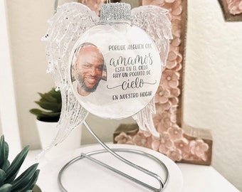 Puede incluir: Una bola de Navidad de cristal transparente con una foto de una persona en su interior, rodeada de alas de ángel decorativas. El adorno tiene purpurina plateada en la parte superior y está suspendido de un soporte plateado. El texto en español dice: "Porque alguien que amamos está en el cielo, hay un poquito de cielo en nuestro hogar."