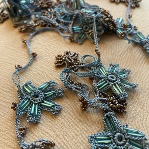 Può includere: Primo piano di gioielli fatti a mano con perline blu e marroni. I gioielli presentano intricati disegni floreali con perline turchesi e marroni. I pezzi sono disposti su una superficie neutra, mostrando la delicata maestria artigianale e la tavolozza dei colori.