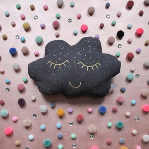 May include: A dark gray cloud-shaped pillow with gold embroidered eyes, a smile, and stars. The pillow is surrounded by colorful pom-poms on a pink background.