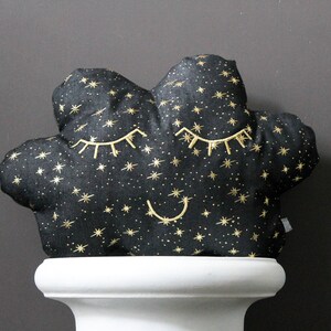 Könnte beinhalten: Ein schwarzes und goldenes Plüschkissen in Wolkenform mit gestickten Augen und einem lächelnden Mund. Das Kissen ist mit goldenen Glitzersternen bedeckt.
