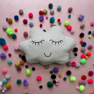 Può includere: Un cuscino bianco a forma di nuvola con un viso sorridente e gli occhi chiusi. Il cuscino è circondato da pompon colorati su uno sfondo rosa.
