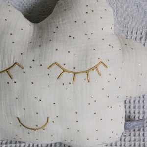 May include: A white cloud-shaped pillow with gold embroidered eyes and a smile. The pillow is covered in small gold dots.