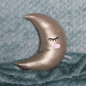 May include: A gold crescent moon-shaped pillow with a smiling face, closed eyes, and pink cheeks. The pillow is lying on a teal fuzzy blanket.