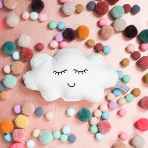 Könnte beinhalten: Ein weißes, wolkenförmiges Kissen mit einem lächelnden Gesicht und geschlossenen Augen. Das Kissen ist von bunten Pompons auf einem rosa Hintergrund umgeben.