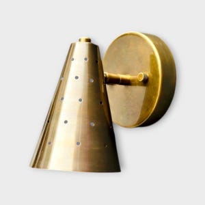 Op de afbeelding: Een messing wandlamp met een conische kap met gelijkmatig verdeelde perforaties. De lamp heeft een cirkelvormige achterplaat en een gebogen arm die de kap met de achterplaat verbindt. De afwerking is een warm, metallic goud.