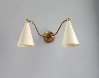 May include: A vintage-style wall sconce with two cream-colored, cone-shaped shades. The shades are connected by curved, brass-toned arms to a circular brass base. The fixture is mounted on a light blue wall, offering a classic lighting design.