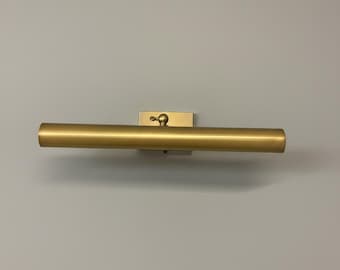 May include: A gold-colored, cylindrical wall-mounted light fixture. The light has a rectangular base and a rounded, horizontal light bar. The fixture is mounted on a gray wall.