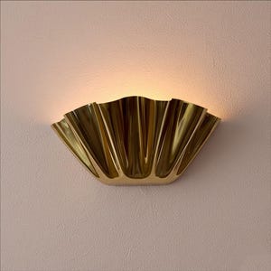 Könnte beinhalten: Goldfarbene Wandleuchte mit einem fächerförmigen Design. Sie ist an einer strukturierten, hellrosa Wand angebracht und strahlt warmes Licht aus. Das Design der Leuchte erinnert an eine Muschel.