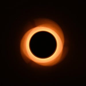 May include: Abstract image of a black circle surrounded by a glowing orange ring, set against a dark background. The ring has a textured appearance, with a subtle spiral effect. The overall effect is reminiscent of a celestial body.