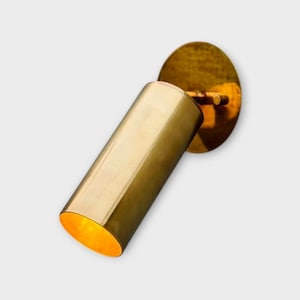 May include: A brass-colored wall-mounted spotlight with a cylindrical body and a circular mounting plate. The light is angled, and the interior of the light is illuminated with a warm glow. The fixture is made of metal.