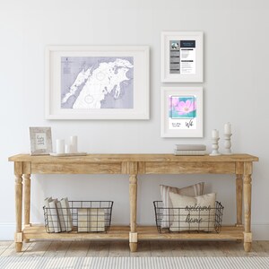 Pode incluir: Uma mesa consola de madeira com duas prateleiras, uma moldura branca com um mapa, uma moldura branca com um desenho floral, uma moldura branca com texto, duas cestas de arame preto e uma almofada com o texto "bem-vindo a casa".