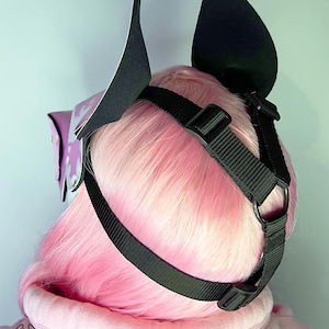 May include: A black and white ear headpiece with a black strap and buckle. The headpiece is worn over pink hair and a pink hoodie. The headpiece has a purple accent with a white design. The text "GRUNGE BUNNY" is visible.