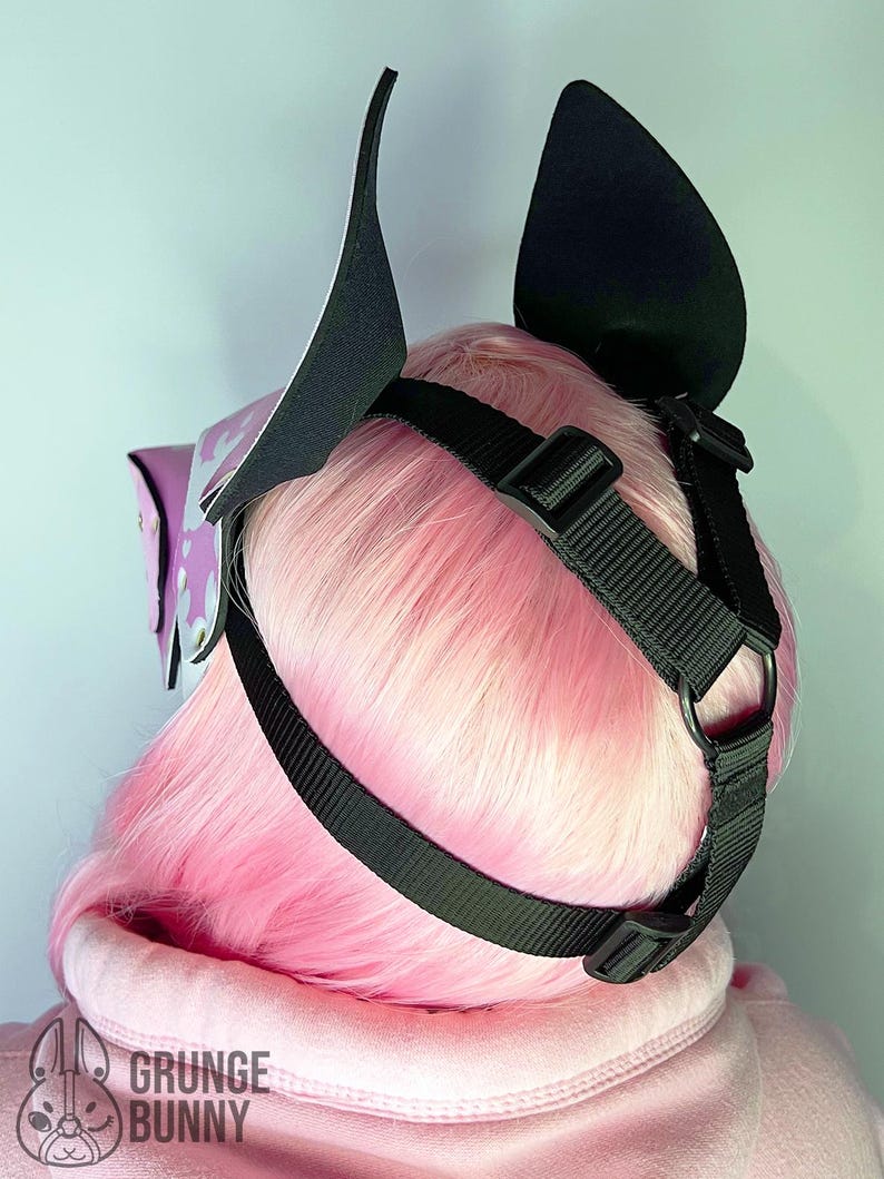 May include: A black and white ear headpiece with a black strap and buckle. The headpiece is worn over pink hair and a pink hoodie. The headpiece has a purple accent with a white design. The text "GRUNGE BUNNY" is visible.