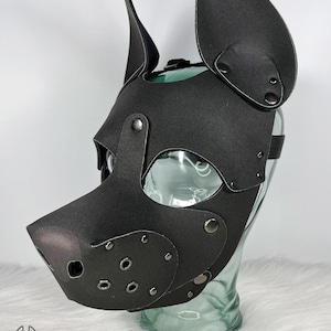 May include: Black pup mask with large, upright ears and a snout. The mask has eye openings and is secured with rivets and a strap. The mask is made of a matte material and is displayed on a clear glass form. The logo "Grunge Bunny" is visible.