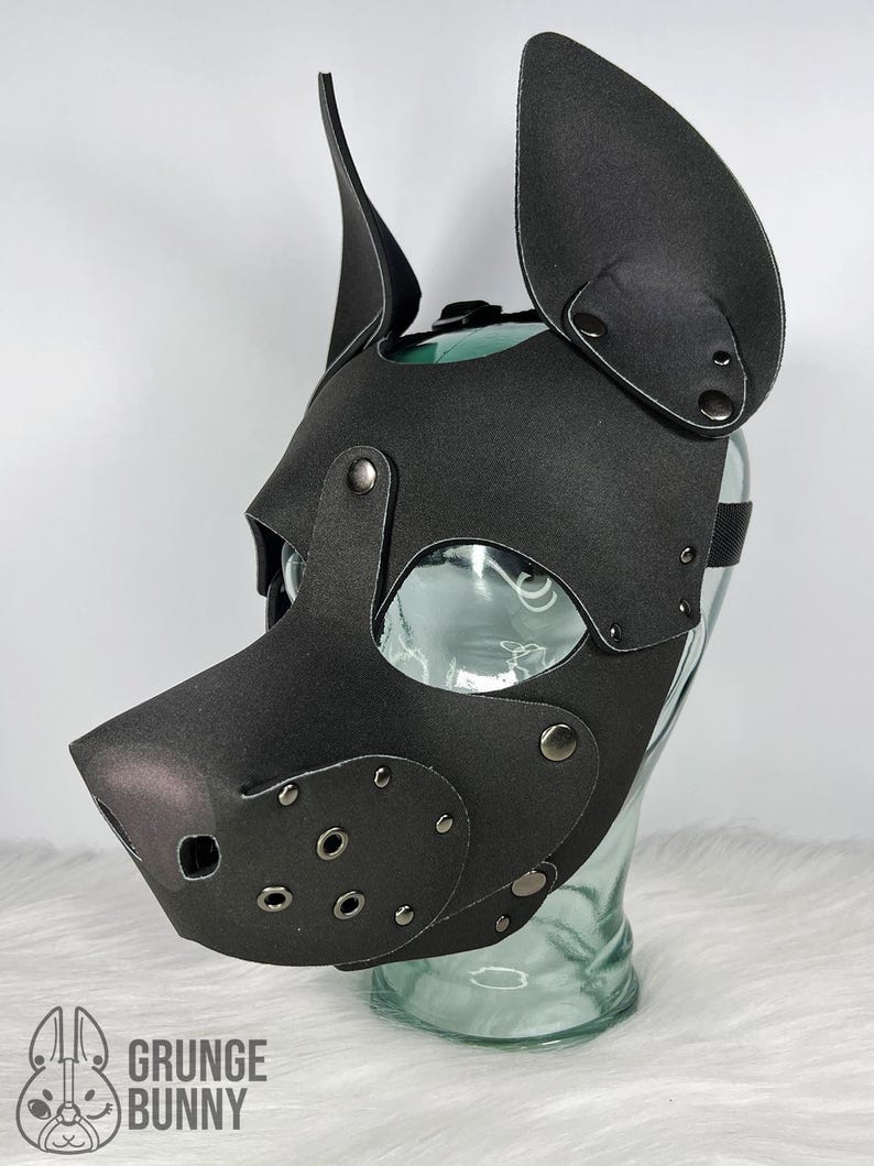 May include: Black pup mask with large, upright ears and a snout. The mask has eye openings and is secured with rivets and a strap. The mask is made of a matte material and is displayed on a clear glass form. The logo "Grunge Bunny" is visible.
