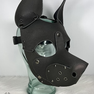 May include: A black pup mask with large, upright ears, eye openings, and a snout. The mask is made of a matte material and features metal rivets and a black adjustable strap. The mask is displayed on a clear glass head form.