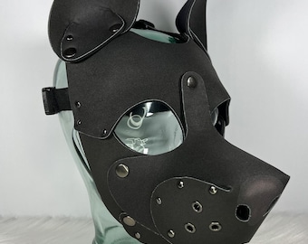 May include: A black pup mask with large, upright ears, eye openings, and a snout. The mask is made of a matte material and features metal rivets and a black adjustable strap. The mask is displayed on a clear glass head form.