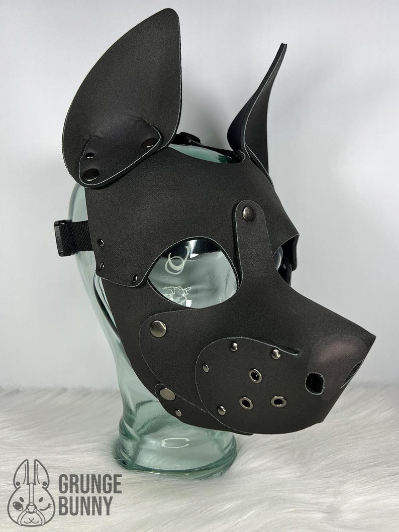 May include: A black pup mask with large, upright ears, eye openings, and a snout. The mask is made of a matte material and features metal rivets and a black adjustable strap. The mask is displayed on a clear glass head form.