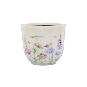 Könnte beinhalten: Ein weißer Keramikübertopf mit facettierter Außenfläche, verziert mit einem zarten botanischen Druck aus violetten, blauen und braunen Blumen, grünem Blattwerk und kleinen goldenen Schmetterlingen sowie einem kleinen blauen Vogel.