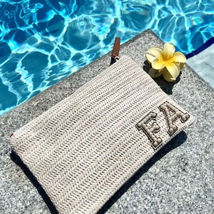 May include: A beige woven clutch bag with the letters "FA" in gold-toned embellishments sits on a stone surface beside a yellow flower. Clear blue water forms the background.