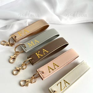 May include: Five personalised keychains in gold, grey, brown, pink, and cream. Each keychain has gold-coloured hardware and gold lettering with initials. The keychains are arranged on a white surface.