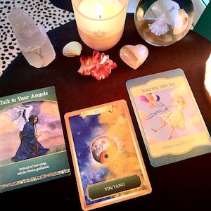 Puede incluir: Tres cartas de oráculo sobre una mesa de madera con una vela, una bola de cristal y cristales. Las cartas son "Habla con tus ángeles", "Yin/Yang" y "Elevarse hacia la alegría".