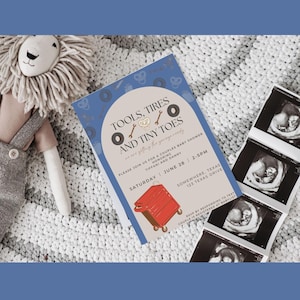 May include: A baby shower invitation with the text "TOOLS, TIRES AND TINY TOES" and a red toolbox graphic. The invitation is next to ultrasound images and a stuffed lion toy on a white and gray blanket.