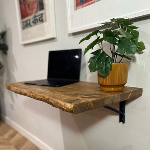 May include: A rustic wooden floating shelf with a laptop and a potted plant on top. The shelf is mounted to a white wall with black metal brackets. The floor is a herringbone pattern.