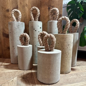 May include: A set of six concrete doorstops with rope handles. The doorstops are various sizes and are arranged on a wooden floor.