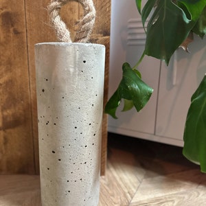 May include: A grey concrete doorstop with a rope handle. The doorstop is sitting on a wooden floor.