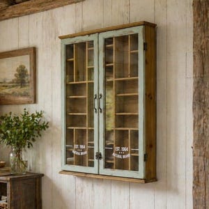 May include: A wall-mounted wooden display cabinet with two glass doors featuring a light blue-green frame. The interior contains multiple small compartments for storage. The glass doors have white text printed on them reading EST. 1964 NATURAL BRAND.
