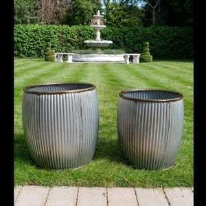 May include: Two cylindrical planters made of corrugated galvanised metal with a dark brown rim. The planters have a vertical ribbed texture and a rounded barrel-like shape, standing side by side on a green lawn in an outdoor garden setting.